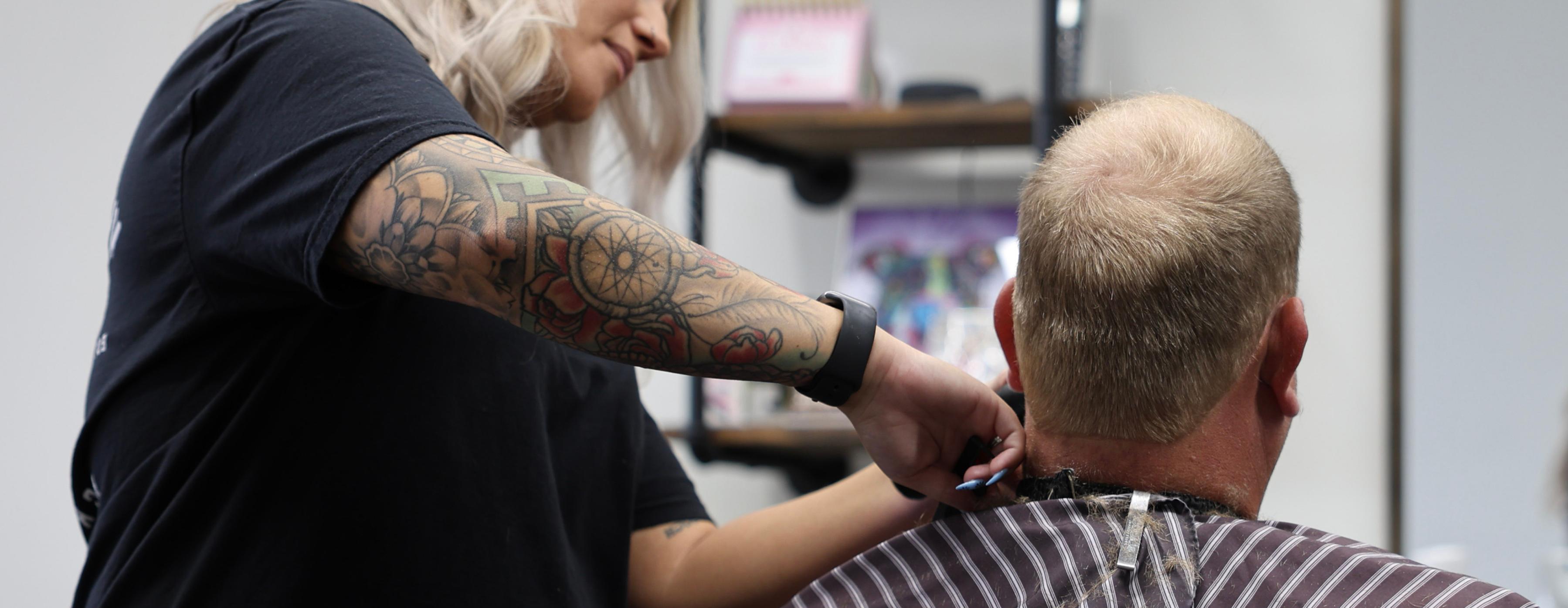 A tattooed hairstylist trims the hair on the back of a mans neck in a salon, with shelves and a striped cape visible in the background.
