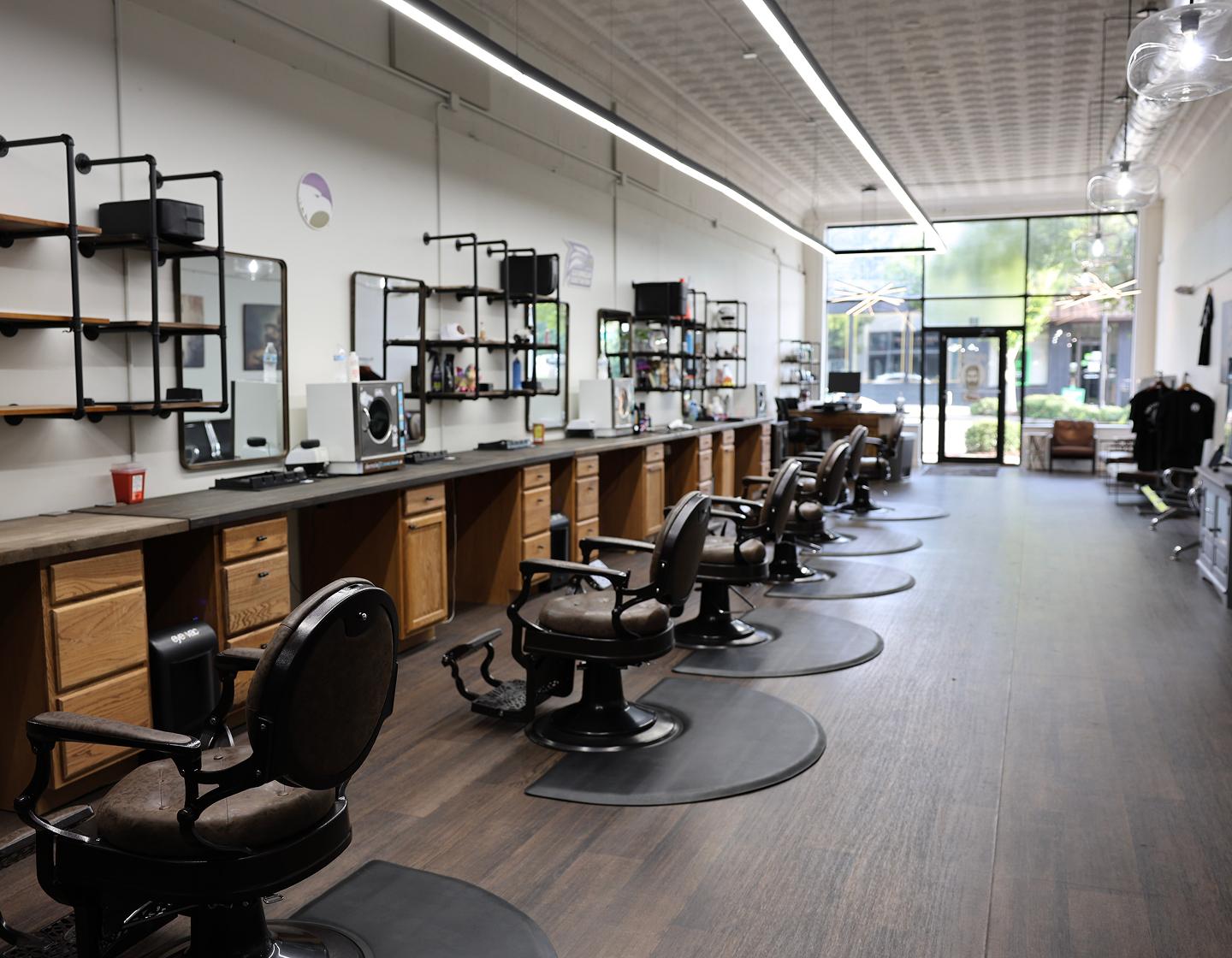 A modern barbershop with empty barber chairs, large mirrors, wooden cabinets, and bright lighting. The shop has a clean, organized appearance with a clear view of the entrance and windows at the far end.