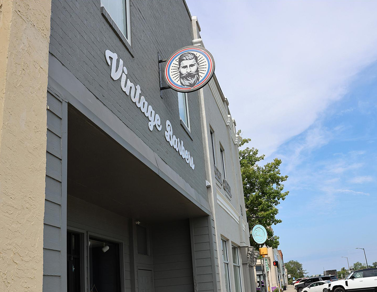 A gray building with a sign reading Vintage Barbers and a circular logo featuring a bearded man. The street outside has trees, parked cars, and other storefronts under a partly cloudy sky.