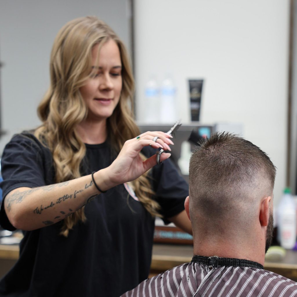 A hairstylist with long, wavy hair is cutting a mans short hair in a salon. The man wears a striped cape and sits facing away from the camera, while the stylist holds scissors above his head.